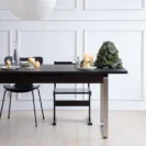 A modern dining table with black chairs is set with candles, plates, a water bottle, and a small decorative Christmas tree. The background features white panelled walls and a light wood floor.