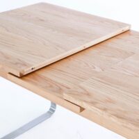 A close-up view of a light wood dining table with an extension leaf partially inserted, showing the mechanism for extending the table. The table has a smooth surface and modern design.
