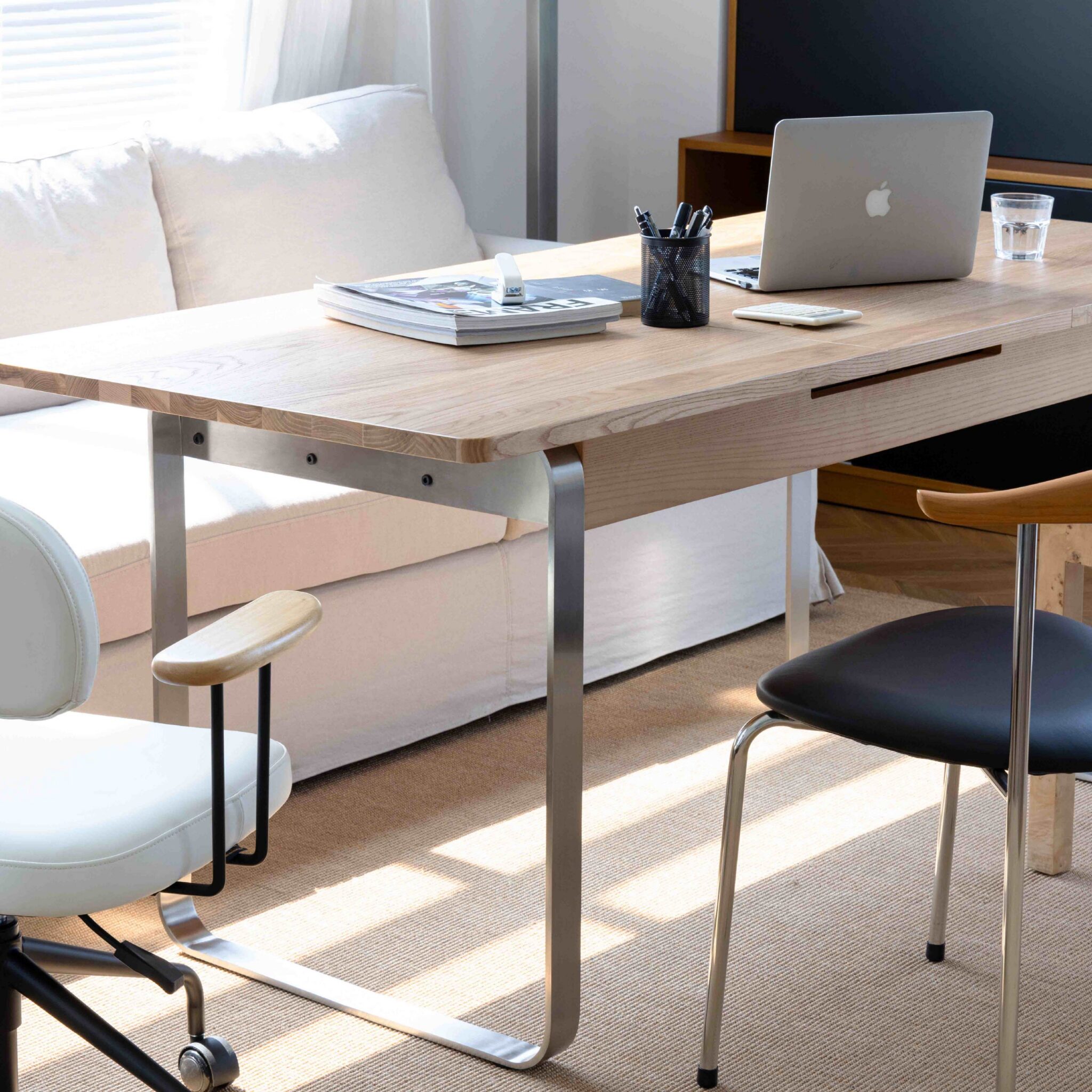 A modern wooden desk with a laptop, magazines, a pen holder, and a glass of water sits in front of a white sofa. Two chairs are placed around the desk in a bright, sunlit room.