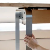 A person’s hand touches a shiny metal table leg that curves upward and attaches to a wooden tabletop with visible screws. The background is softly blurred.