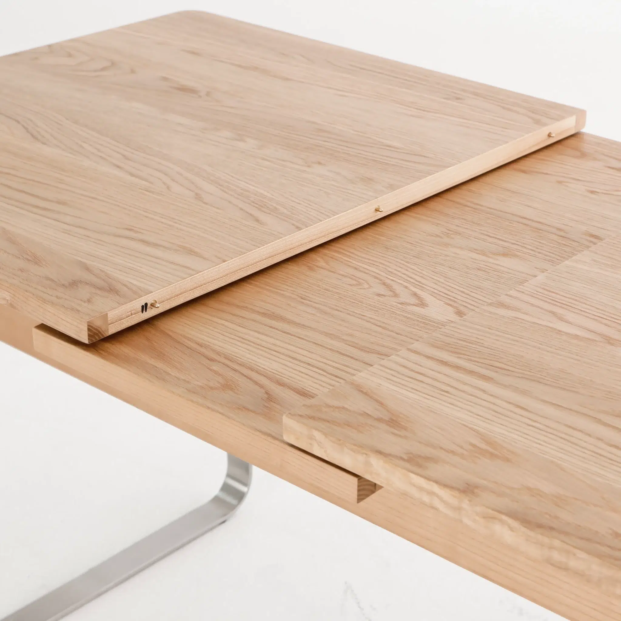 A close-up of a light wood extendable table, partially extended with a leaf insert placed between the two main sections, showing the grain and joinery details.