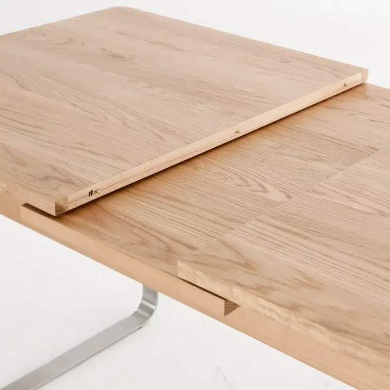 A close-up of a light wood extendable table, partially extended with a leaf insert placed between the two main sections, showing the grain and joinery details.