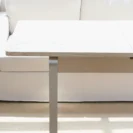 A light wooden table with metal legs is positioned in front of a white sofa on a beige rug, in a brightly lit room.