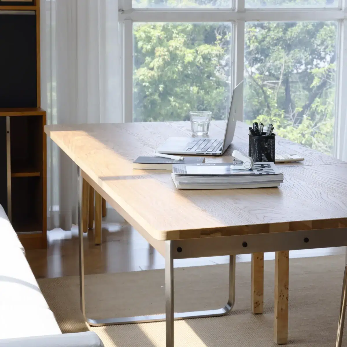 Bauhaus Extendable Dining Table_Natural_Side View A bright workspace with a wooden table, open laptop, glass of water, notebooks, and a holder with pens, set near large windows with sunlight streaming in and trees visible outside.