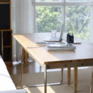 A bright workspace with a wooden table, open laptop, glass of water, notebooks, and a holder with pens, set near large windows with sunlight streaming in and trees visible outside.