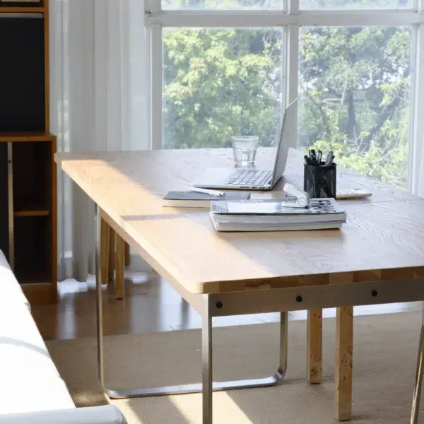 A bright workspace with a wooden table, open laptop, glass of water, notebooks, and a holder with pens, set near large windows with sunlight streaming in and trees visible outside.