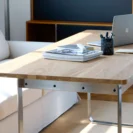 A wooden desk with a metal frame holds a stack of magazines, a tape dispenser, a pen holder with pens, a keyboard, and a laptop. A white sofa and a bookcase are in the background.