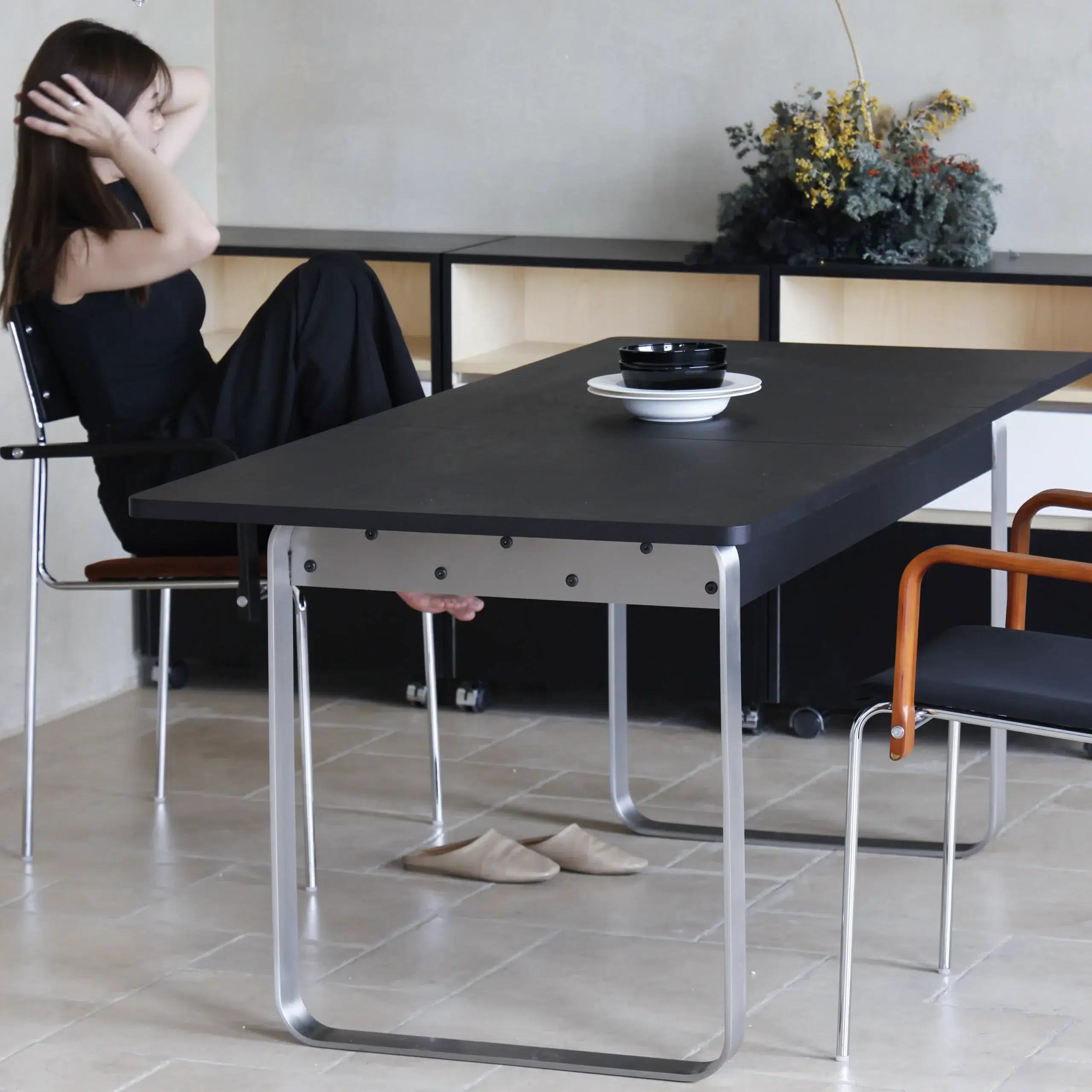 A woman in a black dress sits at a modern black table with metal legs. She adjusts her hair, and a bowl and plate are on the table. Slippers are on the floor, and a plant arrangement is in the background.
