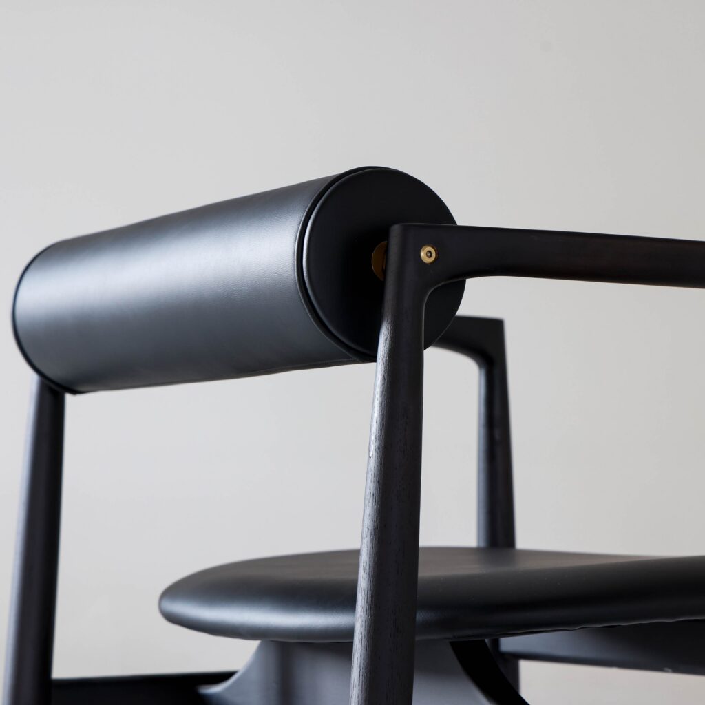 Close-up of a modern black chair with a cylindrical backrest, smooth leather seat, and sleek wooden frame, set against a plain light background.
