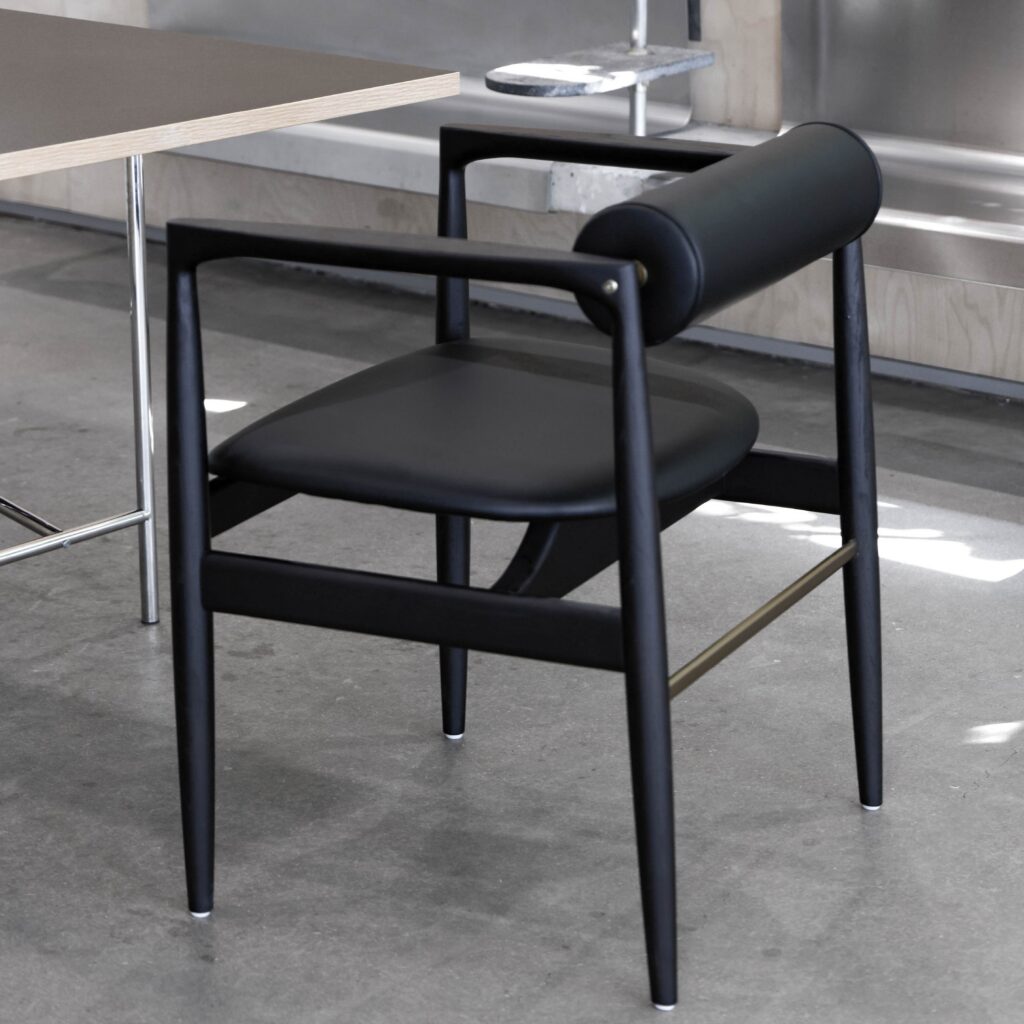 A modern black chair with a cylindrical backrest and slim, angled legs sits on a concrete floor next to a table with a thin wooden top and metal legs.