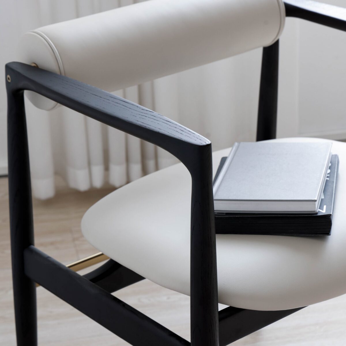 Black Frame + White Seat_C1 A modern chair with black wooden arms and legs, white cushioned seat and backrest. Two closed notebooks are stacked on the seat. Sheer white curtains and light wooden flooring are in the background.