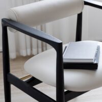A modern chair with black wooden arms and legs, white cushioned seat and backrest. Two closed notebooks are stacked on the seat. Sheer white curtains and light wooden flooring are in the background.