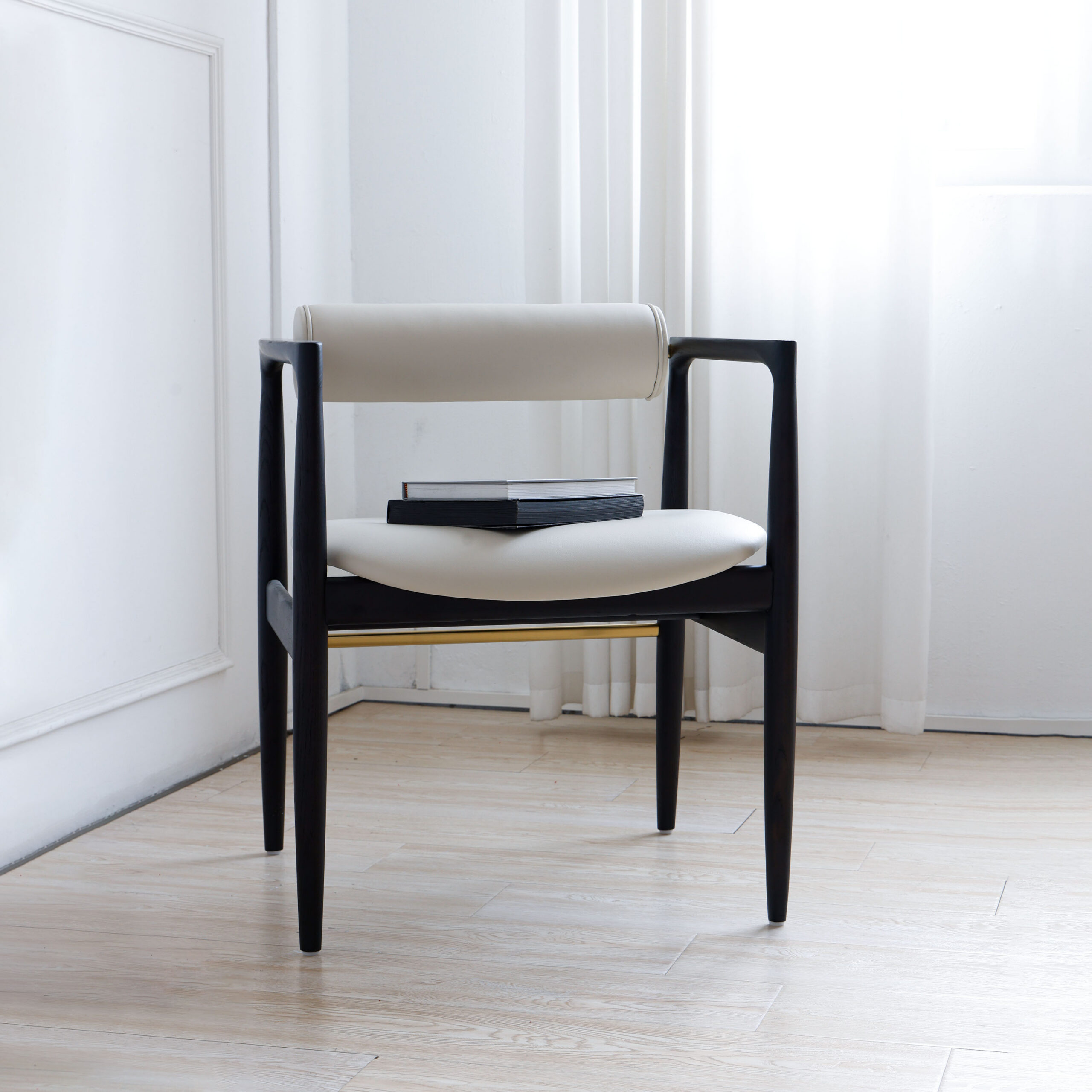 A modern chair with black wooden frame and white cushioned seat and backrest, placed on a light wooden floor near a window with sheer white curtains. Two closed black books rest on the seat.