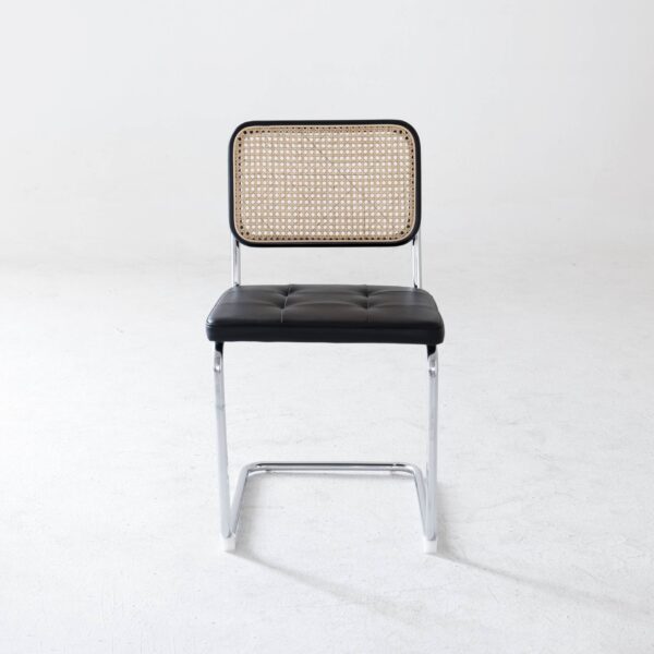 A modern chair with a chrome frame, black cushioned seat, and a rectangular cane backrest, positioned on a plain white floor with a light grey wall in the background.