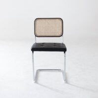 A modern chair with a chrome frame, black cushioned seat, and a rectangular cane backrest, positioned on a plain white floor with a light grey wall in the background.