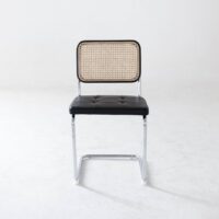 A modern chair with a chrome frame, black cushioned seat, and a rectangular cane backrest, positioned on a plain white floor with a light grey wall in the background.