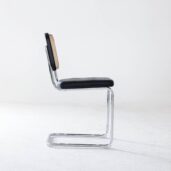 A modern cantilever chair with a chrome metal frame, a black cushioned seat, and a woven cane backrest, shown in profile against a plain white background.