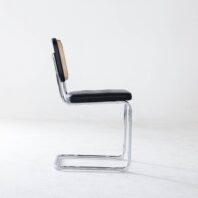 A modern cantilever chair with a chrome metal frame, a black cushioned seat, and a woven cane backrest, shown in profile against a plain white background.