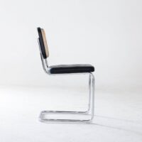 A modern cantilever chair with a chrome metal frame, a black cushioned seat, and a woven cane backrest, shown in profile against a plain white background.