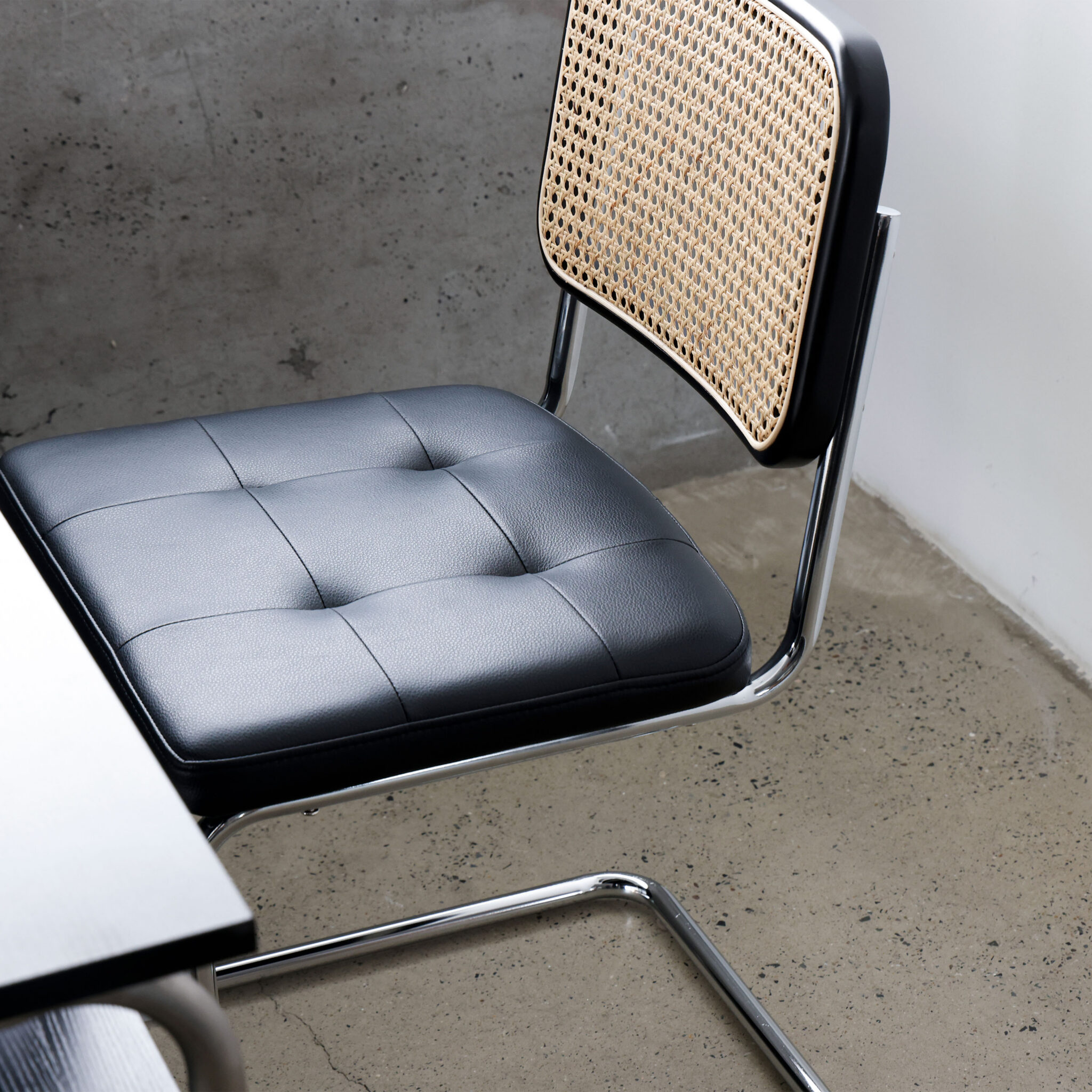 A modern chair with a chrome frame, black cushioned seat, and a woven cane backrest sits next to a black table on a polished concrete floor.