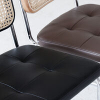 Close-up of three modern chairs with chrome legs; one has a woven cane backrest and brown cushion, the other two have tufted cushions in black and brown faux leather. All are placed on a light-coloured floor.