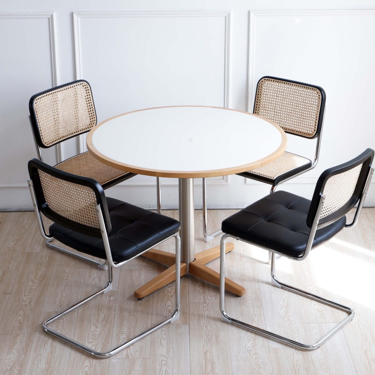 Black Frame_Chrome_Black Leather_C9 A round white table with a wooden edge and base is surrounded by four modern chairs with black cushions, metal frames, and woven cane backs, set on a light wood floor against a white wall.