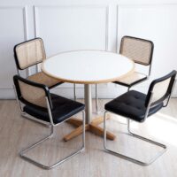 A round white table with a wooden edge and base is surrounded by four modern chairs with black cushions, metal frames, and woven cane backs, set on a light wood floor against a white wall.