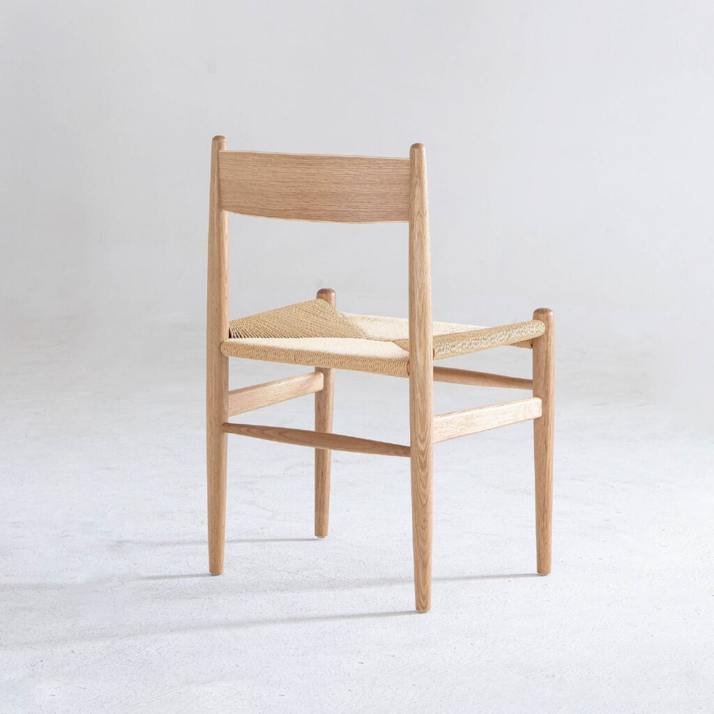A minimalist wooden chair with a woven straw seat, viewed from an angle showing the back and one side, placed on a plain light-coloured floor with a neutral background.