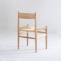 A minimalist wooden chair with a woven straw seat, viewed from an angle showing the back and one side, placed on a plain light-coloured floor with a neutral background.