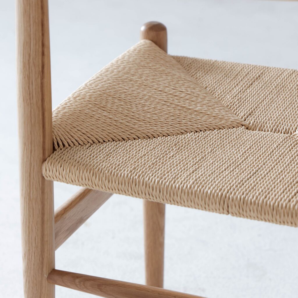 Close-up of a wooden chair with a woven natural fibre seat, showcasing the intricate, diagonal pattern and smooth, light-coloured wood frame. The background is out of focus.
