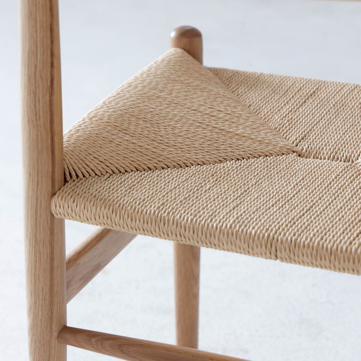 CH36 Chair_B4 Close-up of a wooden chair with a woven natural fibre seat, showcasing the intricate, diagonal pattern and smooth, light-coloured wood frame. The background is out of focus.