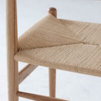 Close-up of a wooden chair with a woven natural fibre seat, showcasing the intricate, diagonal pattern and smooth, light-coloured wood frame. The background is out of focus.
