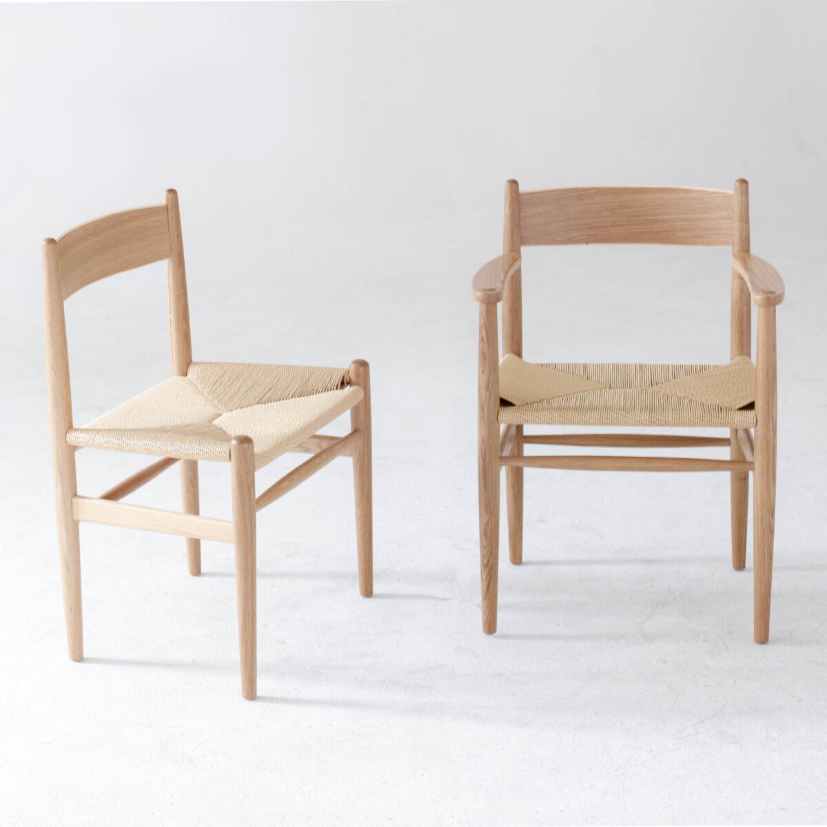 CH36 Chair_B5 Two wooden chairs with light-coloured woven seats are placed side by side on a light, textured floor. One chair has armrests whilst the other does not. Both have simple, minimalist designs.