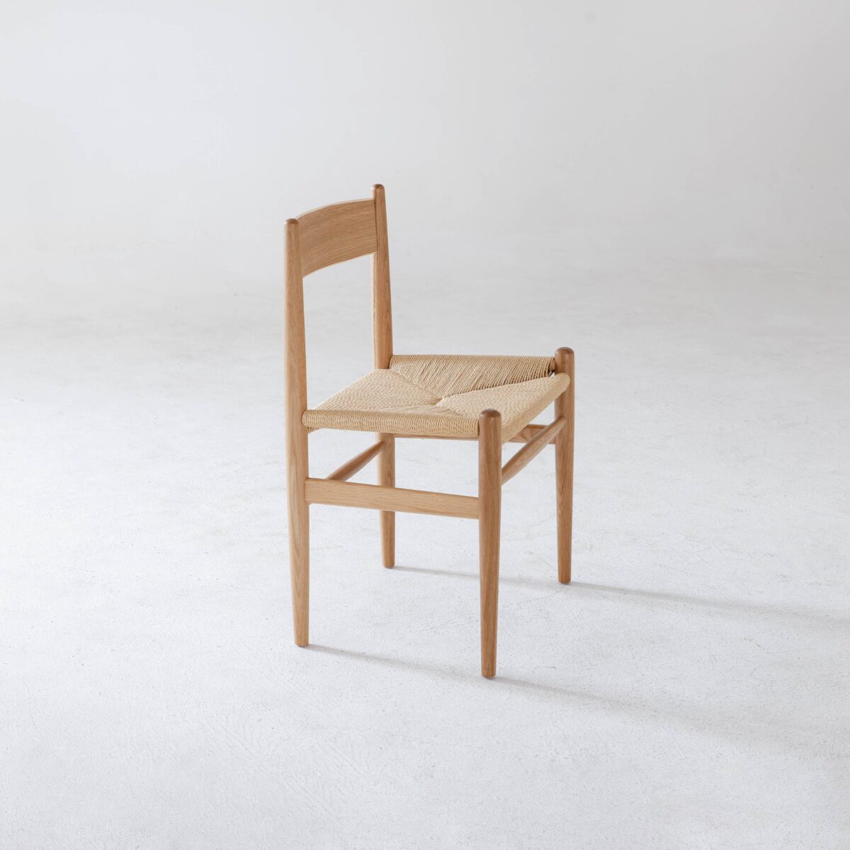 CH36 Chair_B6 A simple wooden chair with a woven straw seat, angled slightly to the left, sits on a plain white floor against a minimal, light background.
