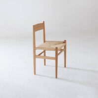 A simple wooden chair with a woven straw seat, angled slightly to the left, sits on a plain white floor against a minimal, light background.