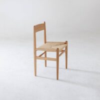 A simple wooden chair with a woven straw seat, angled slightly to the left, sits on a plain white floor against a minimal, light background.