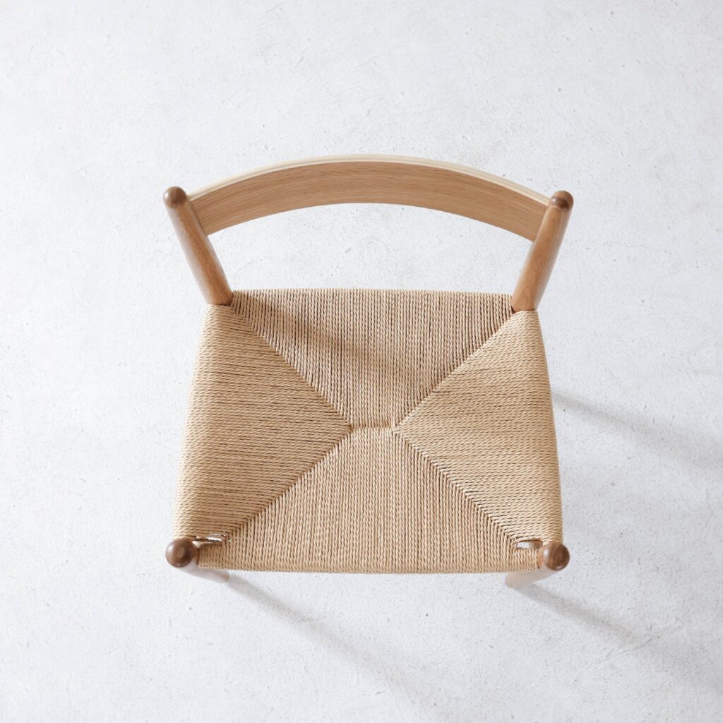 Top view of a wooden chair with a curved backrest and a woven, natural fibre seat, placed on a light, textured surface.