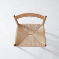 Top view of a wooden chair with a curved backrest and a woven, natural fibre seat, placed on a light, textured surface.