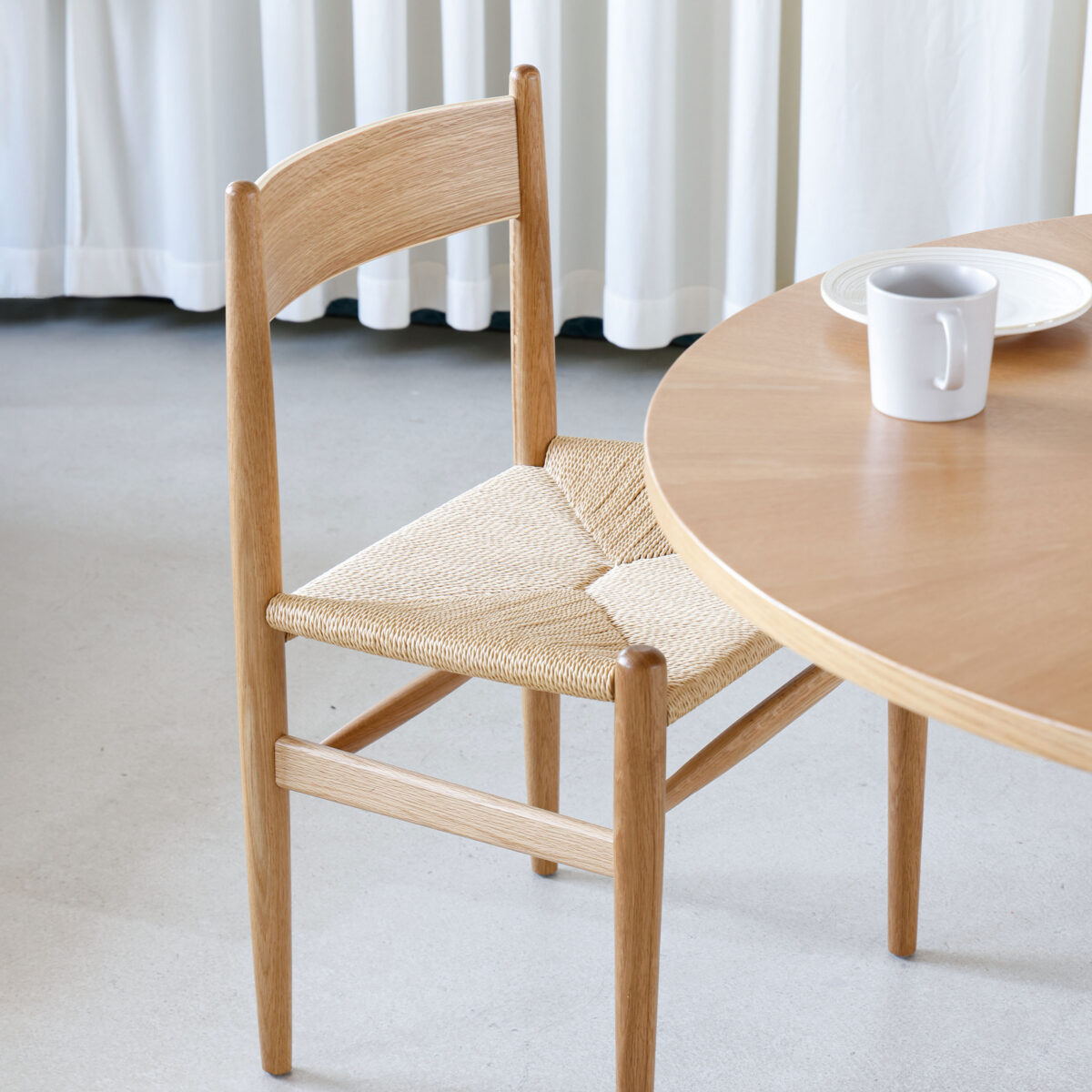 CH36 Chair_C2 A wooden chair with a woven seat is positioned next to a round wooden table. On the table, there is a white mug and a white plate. White curtains hang in the background.