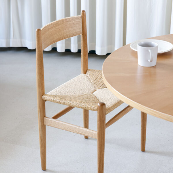 A wooden chair with a woven seat is positioned next to a round wooden table. On the table, there is a white mug and a white plate. White curtains hang in the background.