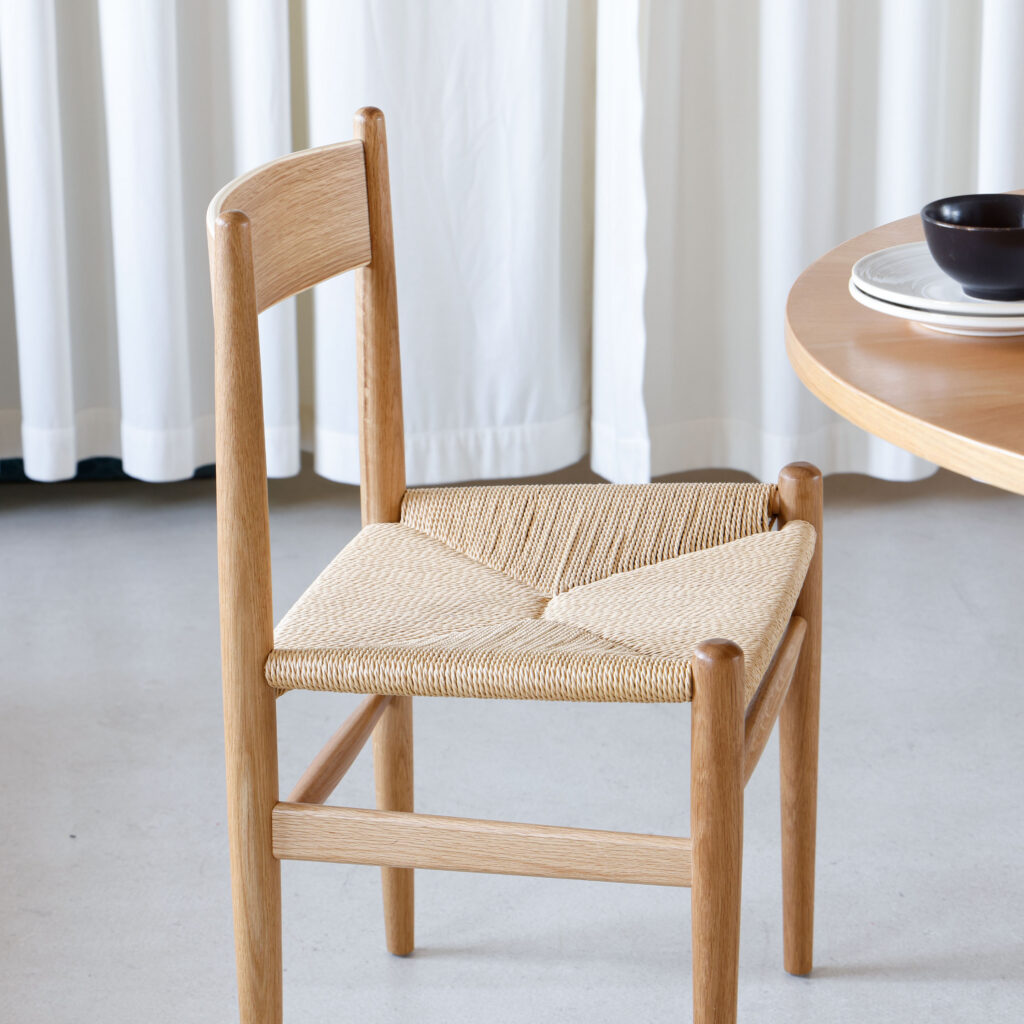 A wooden chair with a woven seat is placed next to a round wooden table set with plates and a bowl. White curtains hang in the background, and the setting has a minimal, modern look.