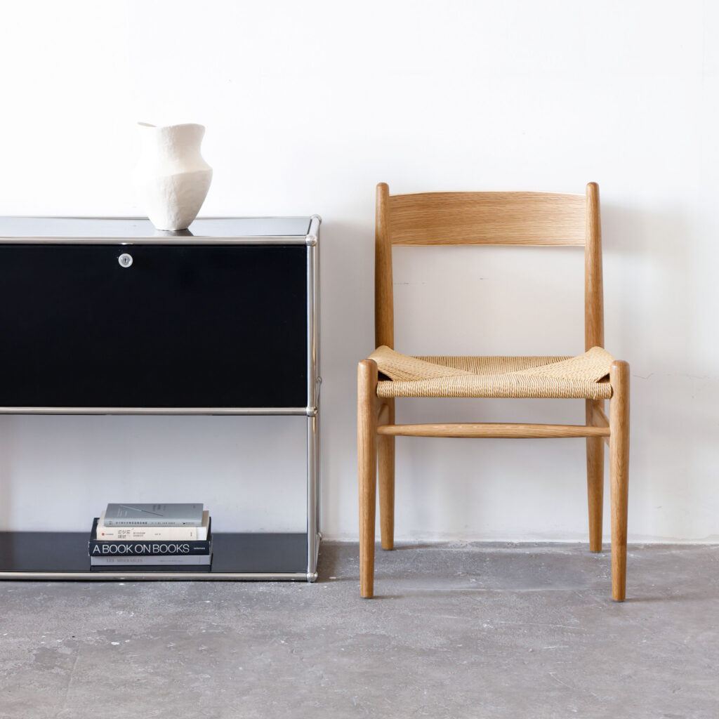 A wooden chair with a woven seat is placed next to a black metal cupboard. On top of the cupboard is a white ceramic vase, and two stacked books are on the lower shelf. The setting is minimal, with a plain white wall and grey floor.