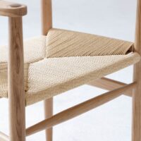 Close-up of a wooden chair featuring a woven natural fibre seat with a geometric pattern, highlighting the texture and craftsmanship of the materials. The background is a plain, light surface.