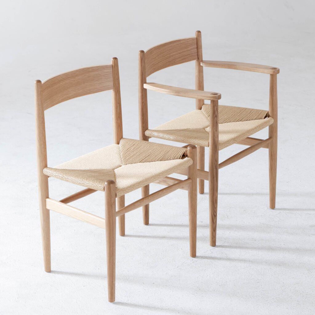 Two wooden chairs with woven seats are positioned side by side on a light surface. One chair has armrests, and the other does not. Both have simple, minimalist designs with natural wood finishes.