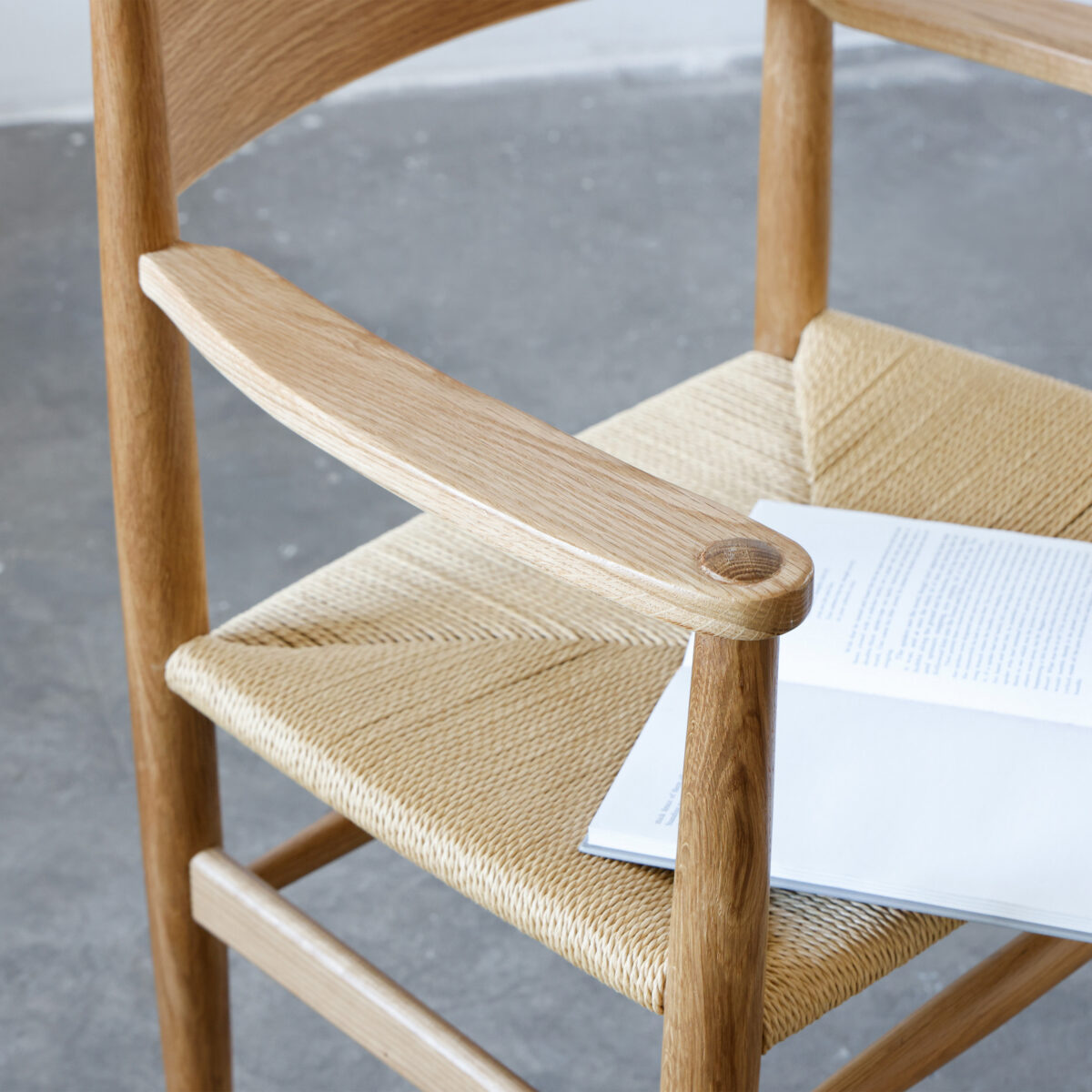 CH37_C2 A close-up of a wooden chair with woven seat, featuring a light wood finish. An open book with printed text rests partially on the seat. The background is a grey concrete floor.