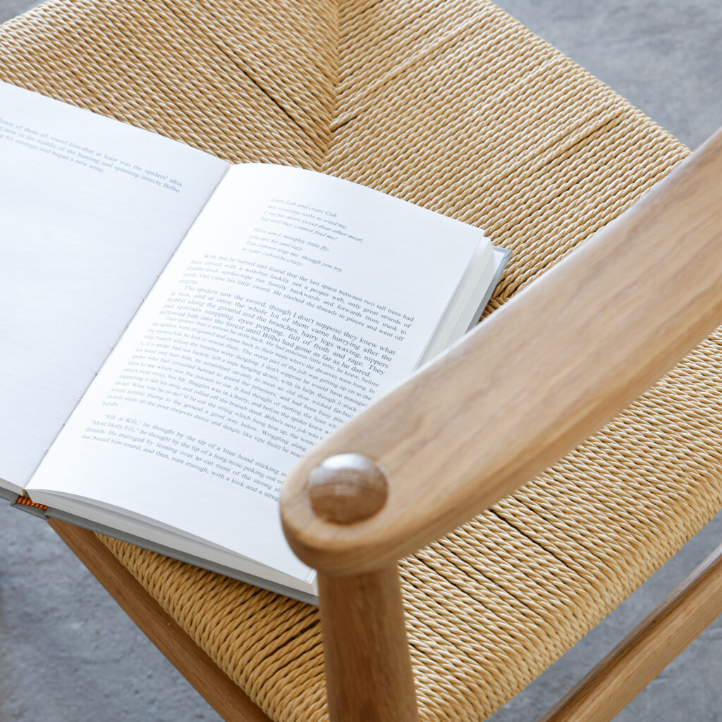 An open book rests on the woven seat of a wooden chair, viewed from above. The chair has a natural finish and is placed on a grey floor.