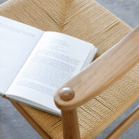 An open book rests on the woven seat of a wooden chair, viewed from above. The chair has a natural finish and is placed on a grey floor.