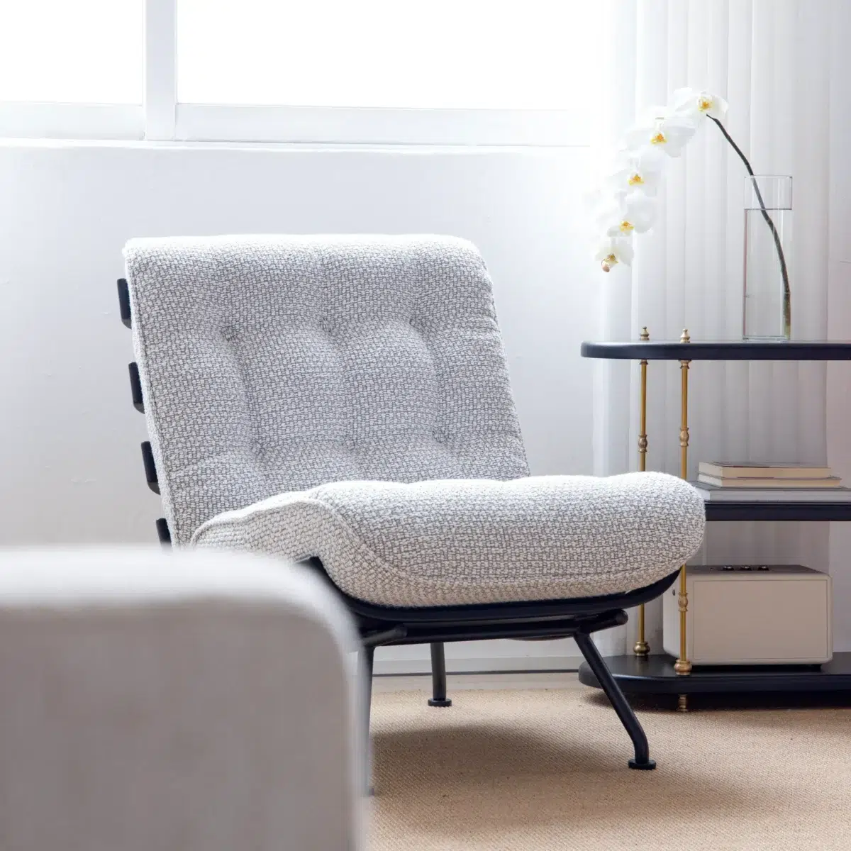 Costela Chair_Front View A modern, textured, light grey lounge chair sits in a bright room near a small black and gold side table holding a white orchid in a glass vase. Light filters in through a large window behind the chair.