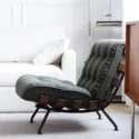 A modern, tufted lounge chair with dark upholstery and a black metal frame sits on a light-coloured rug in a bright living room with a white sofa and minimalist décor.