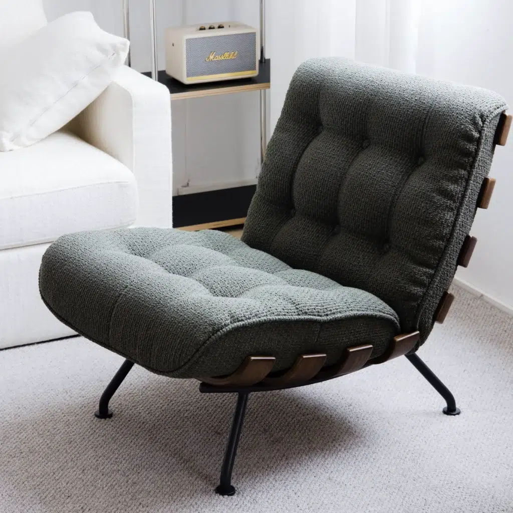 A modern lounge chair with dark green tufted upholstery, black legs, and wooden accents sits next to a white sofa and a shelf holding a beige Marshall speaker in a bright, carpeted room.